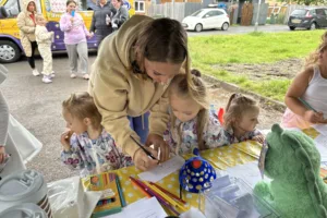 A woman helps two young girls draw at an outdoor community event.