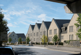 Computer generated image of a row of modern stone buildings with pitched roofs along a street, with trees, a stone wall, and people walking under a clear blue sky