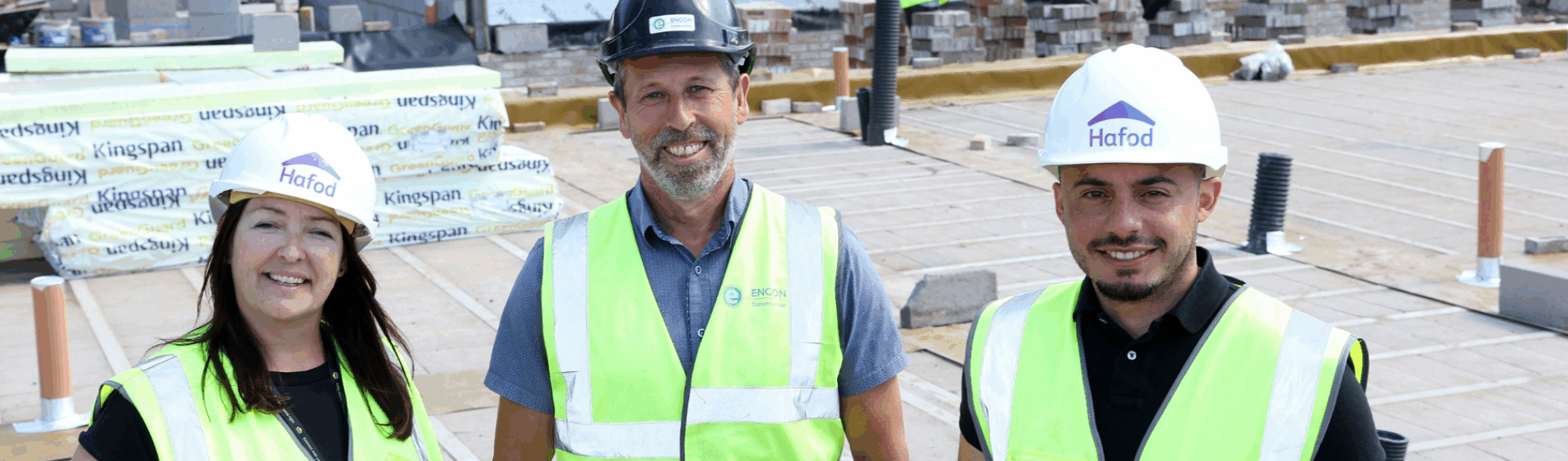 Three people wearing high-visibility vests and hard hats standing on a construction site with building materials and partially built walls in the background