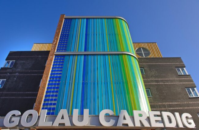 Exterior of the Golau Caredig building, featuring a curved facade with blue and green panels, and silver signage.