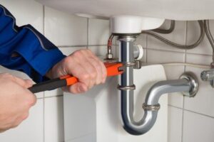 Plumber using a wrench to repair the chrome pipework beneath a white sink.