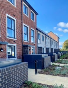 A row of modern terraced houses with a mix of red and light grey brick exteriors. Each house has large rectangular windows and black front doors, with small gated front patios bordered by low brick walls.