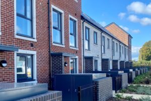 A row of modern terraced houses with a mix of red and light grey brick exteriors. Each house has large rectangular windows and black front doors, with small gated front patios bordered by low brick walls.
