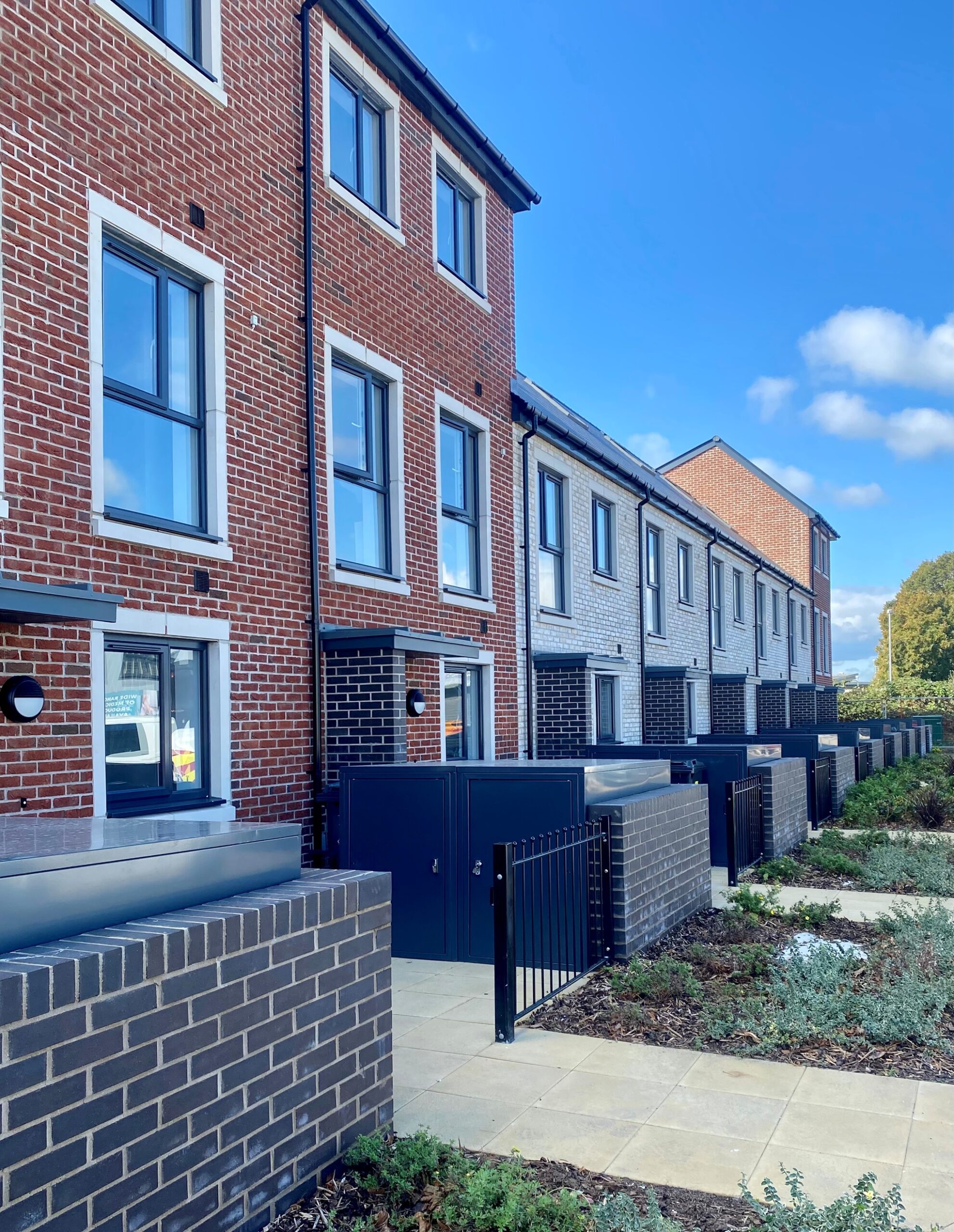 A row of modern terraced houses with a mix of red and light grey brick exteriors. Each house has large rectangular windows and black front doors, with small gated front patios bordered by low brick walls.