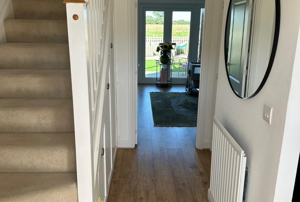 Entrance hallway showing the staircase with banister and spindles to the first floor. Doorway to living room is shown as well as some of the room. Circular mirror displayed on hallway wall.