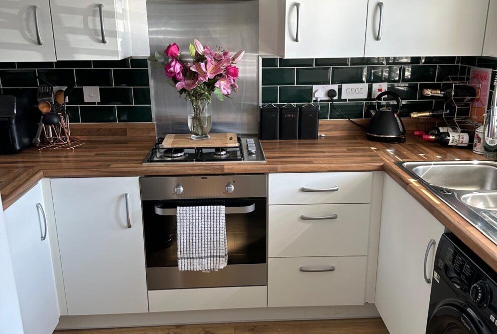 Modern cream kitchen with wood-effect worktops. Green brick-style tiling. Wood-effect flooring. Includes intergral oven and hob, fridge-freezer and washing machine.