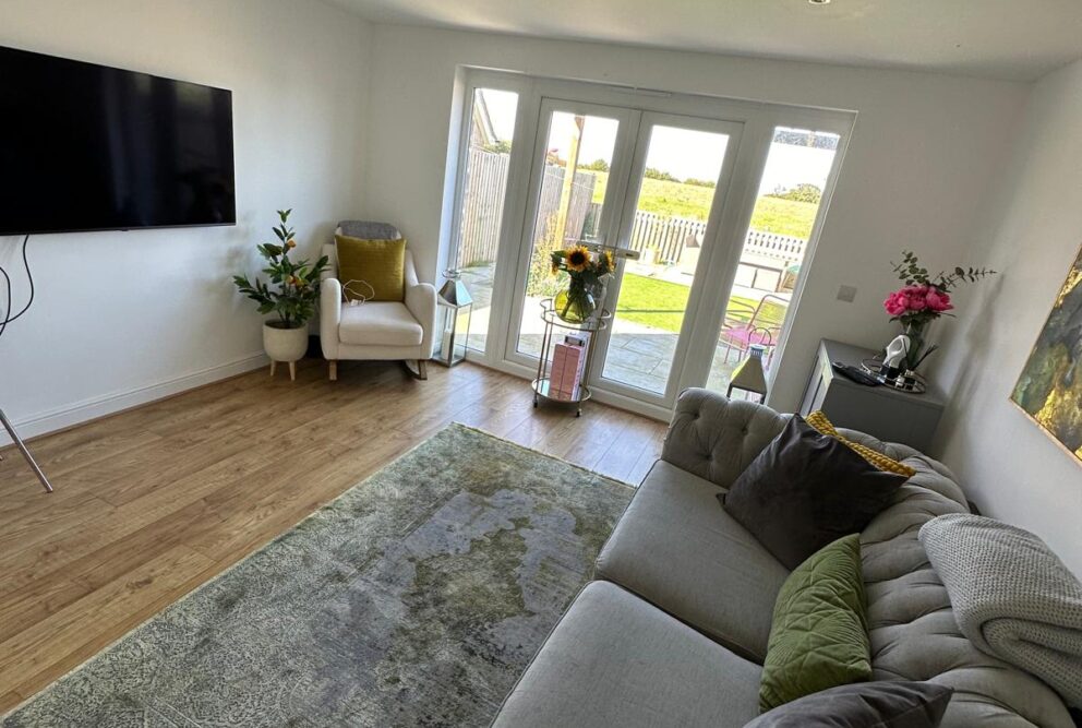 Living room with a grey sofa, white chair, grey patterned rug and television mounted on the wall. The room also has some lovely floral displays and artwork on the wall. Patio doors to access the rear garden.