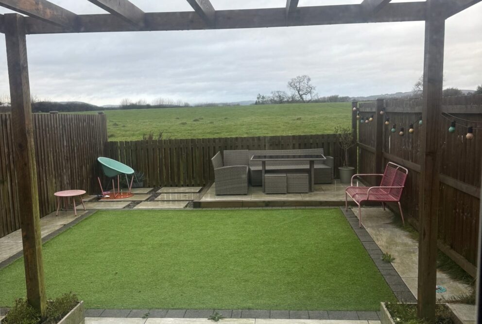 Rear garden view with a pagoda over the patio area, followed by a lawned garden and decked seating area. Views of field behind.