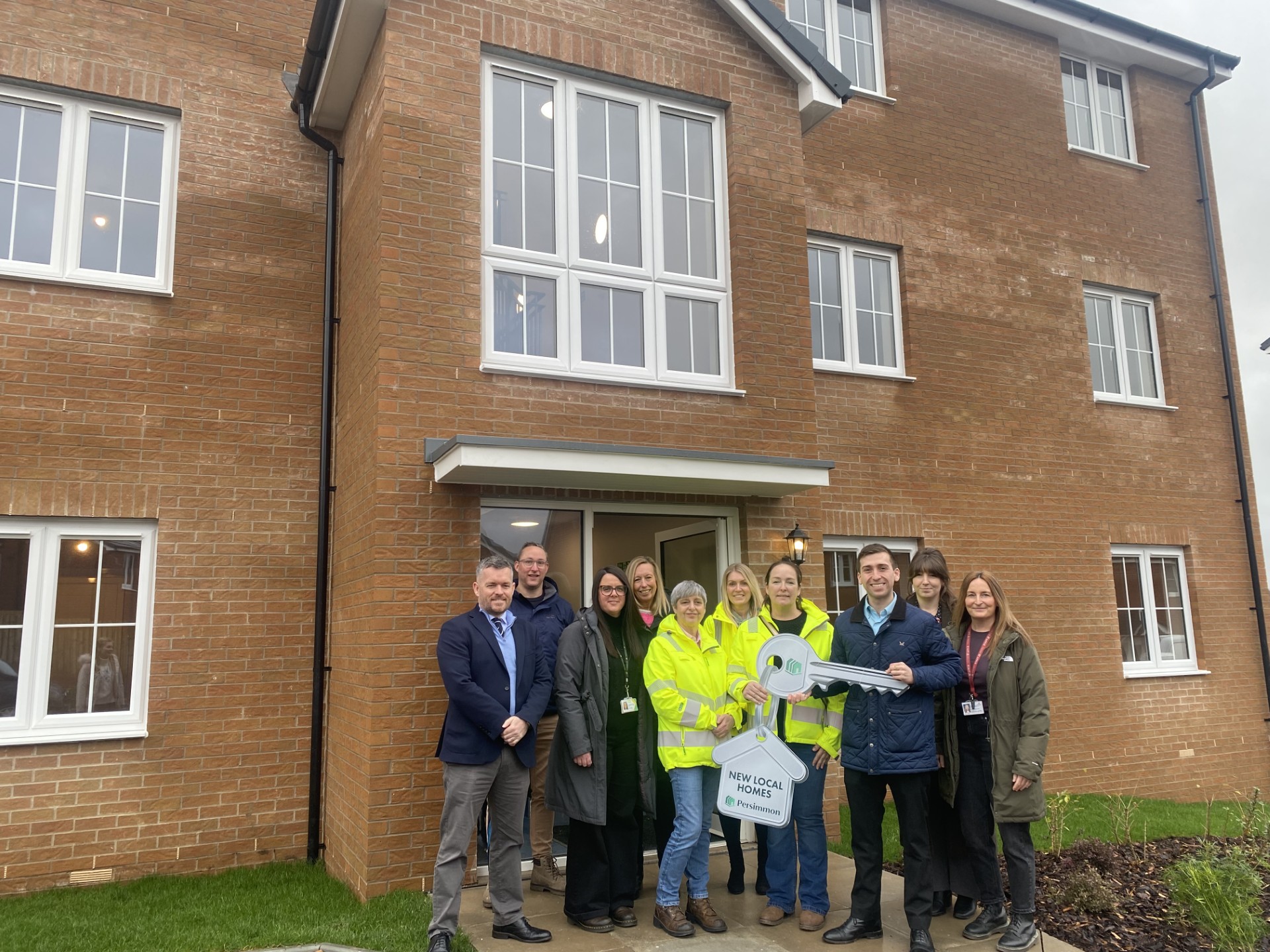 Group of people smiling, one holding a large key, stood in front of a new block of apartments
