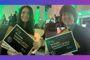 Two people seated indoors at an event venue, each holding a sign that reads “We’ve been shortlisted – Environment Category” for the Travis Perkins Better Together Awards. The background shows tables, chairs, and green event lighting.