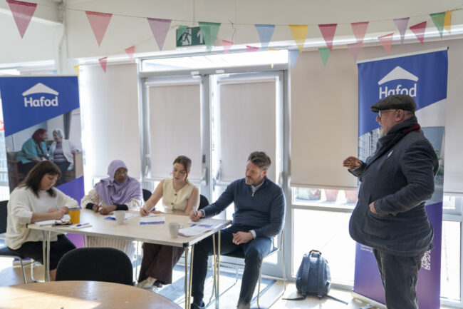 A group of people at a desk and one person standing up talking to them in a room