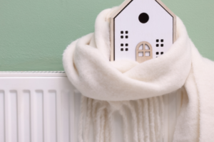 Picture of a small wooden house wrapped in a white scarf sitting on a radiator against a green wall.