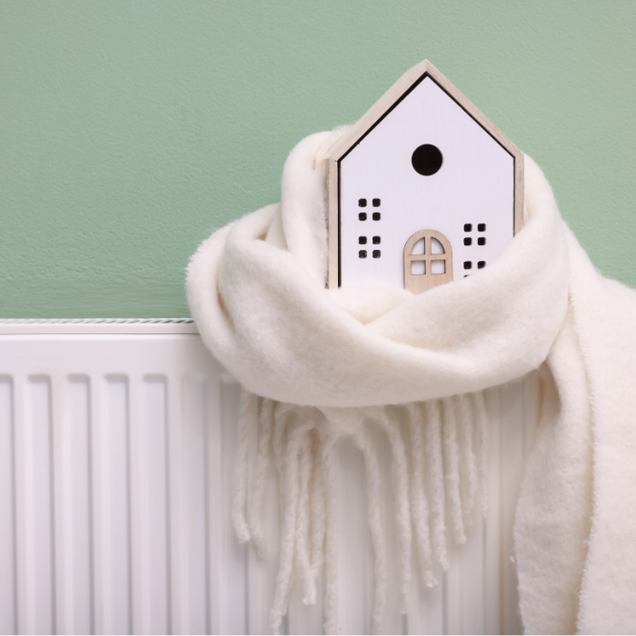 Picture of a small wooden house wrapped in a white scarf sitting on a radiator against a green wall.